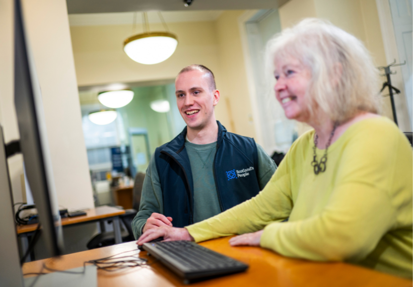 A woman and man seated at a desk, smiling while looking at a computer screen 
